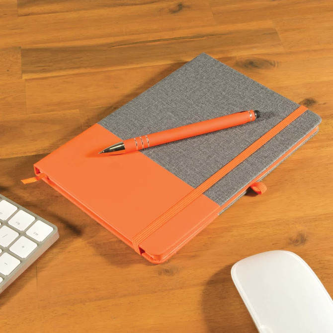 A grey notebook with an orange cover and an orange pen placed on a wooden desk beside a keyboard and mouse.