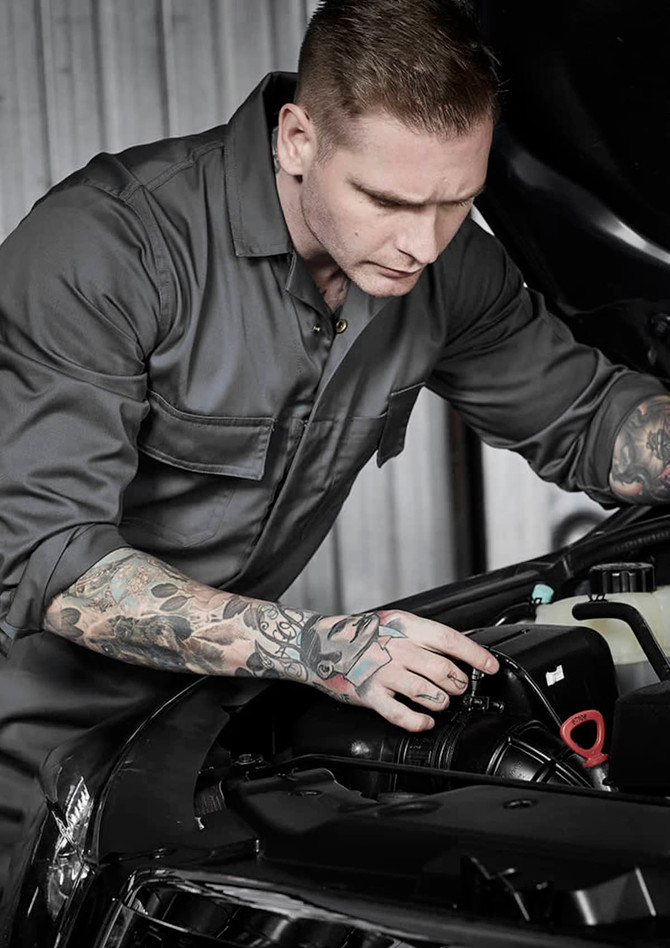 A man in a grey service overall works on a car engine, showcasing detailed tattoos on his arms. The image features a logo on the overall.