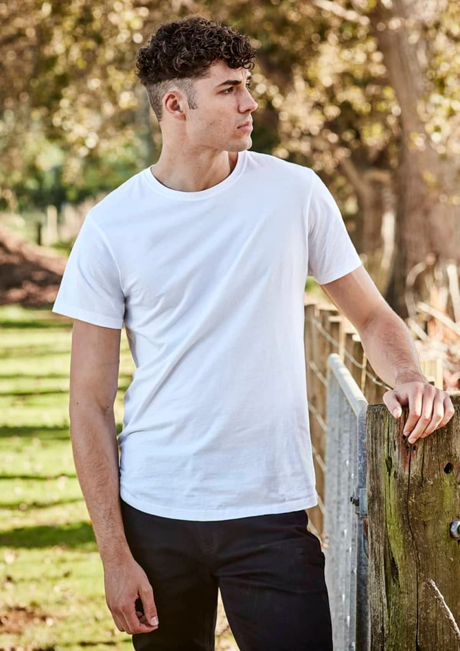 A white men's T-shirt is worn by a model standing beside a wooden fence in an outdoor setting.
