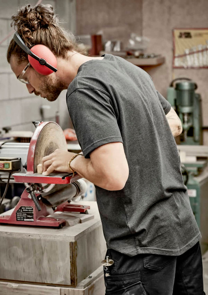 A man wearing a grey T-shirt works on a machine in a workshop, with safety glasses and earmuffs.