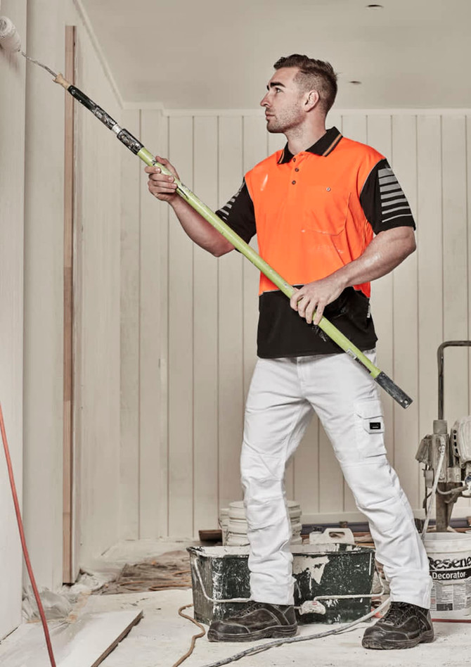 A man in an orange and black short sleeve polo shirt uses a roller to paint a wall. The background has painting equipment.