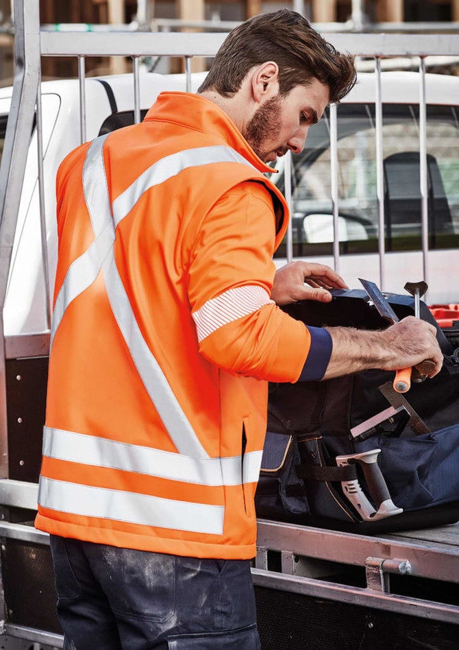 An orange unisex softshell jacket with reflective strips worn by a person at a worksite, with tools nearby.