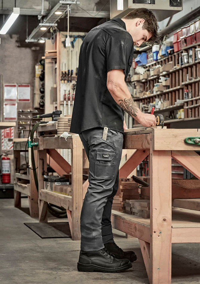 A man in black cargo pants and a black shirt standing at a workbench in a workshop, focused on a task.