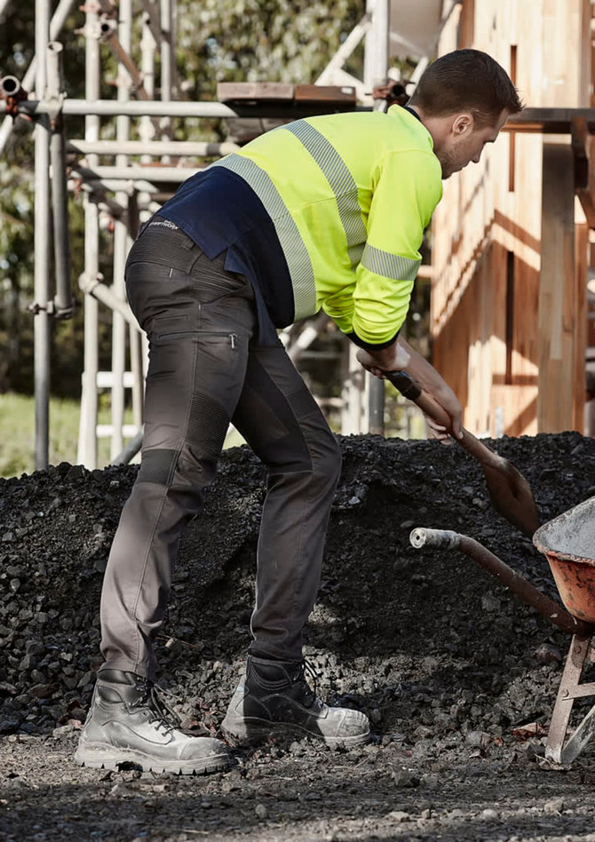 A man in high-visibility workwear and black stretch pants shovels gravel at a construction site.