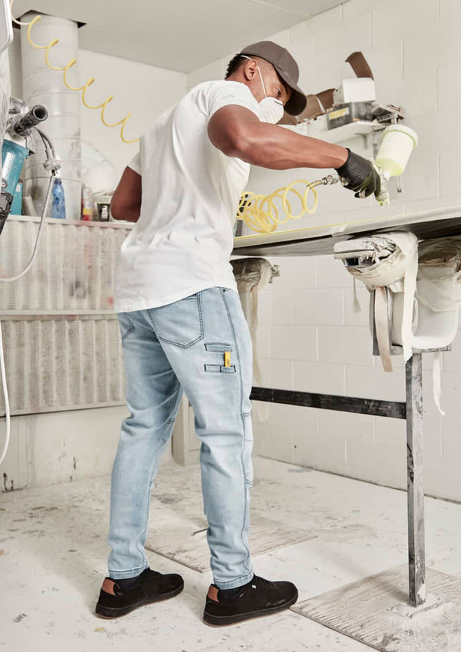 A man wearing a white shirt and grey streetwear stretch jeans, painting in a workshop setting.
