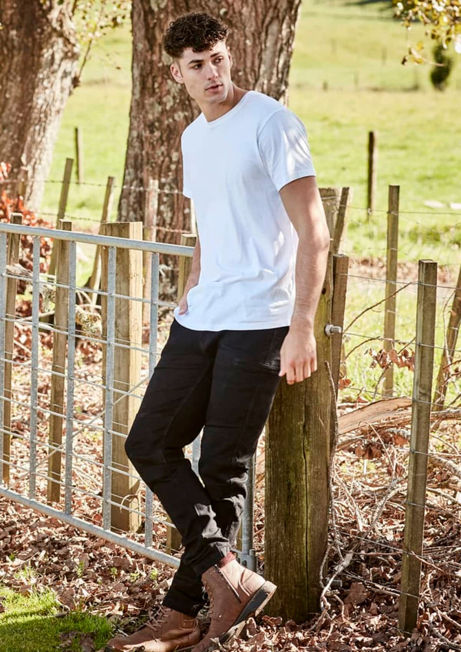 A person wearing black stretch jeans and a white t-shirt stands by a wooden fence in a grassy area.