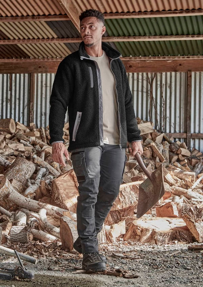 A model wearing black work pants and a black fleece jacket stands outdoors, with stacks of timber in the background.