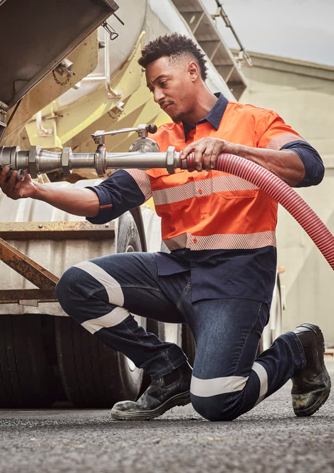 A worker in orange and navy striped attire kneels beside a tanker, wearing Mens Bio Motion Taped Stretch Jeans.