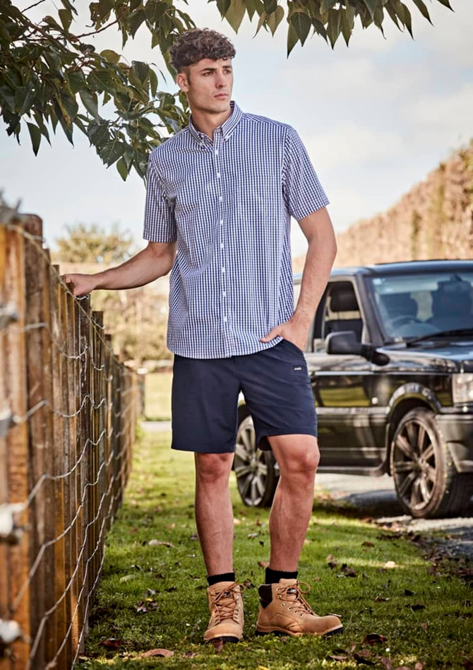 A man wearing a short-sleeve checked shirt and navy lightweight outdoor shorts, standing by a fence.