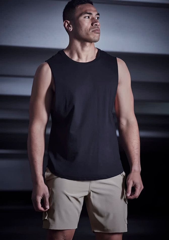 A man wearing a black sleeveless top and beige sports shorts stands in a dimly lit setting. The shorts feature a logo.