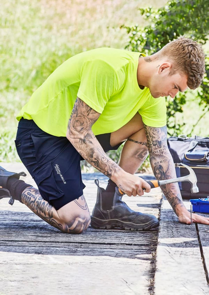 A person kneeling outdoors in bright yellow clothing and dark blue sports shorts, using a saw on a wooden surface.