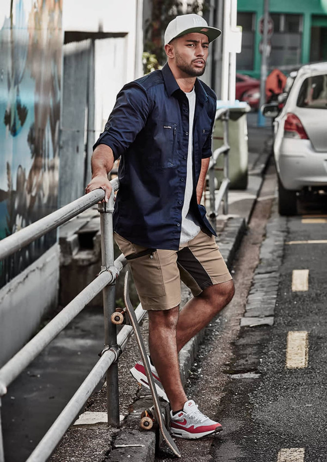 A man in a navy shirt and khaki shorts stands by a railing, with a skateboard and cars in the background.