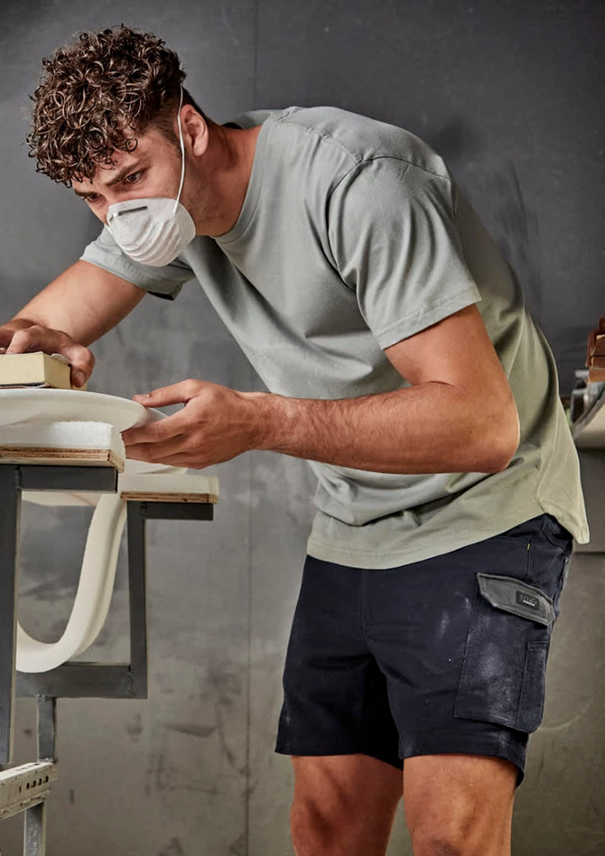 A man wearing a grey t-shirt and navy shorts, working on a project with a mask on in an indoor workspace.