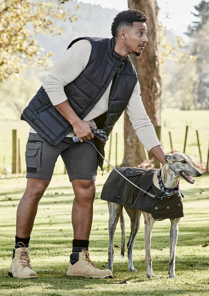 A man wearing a black vest and grey shorts stands with a leashed greyhound in a grassy park.