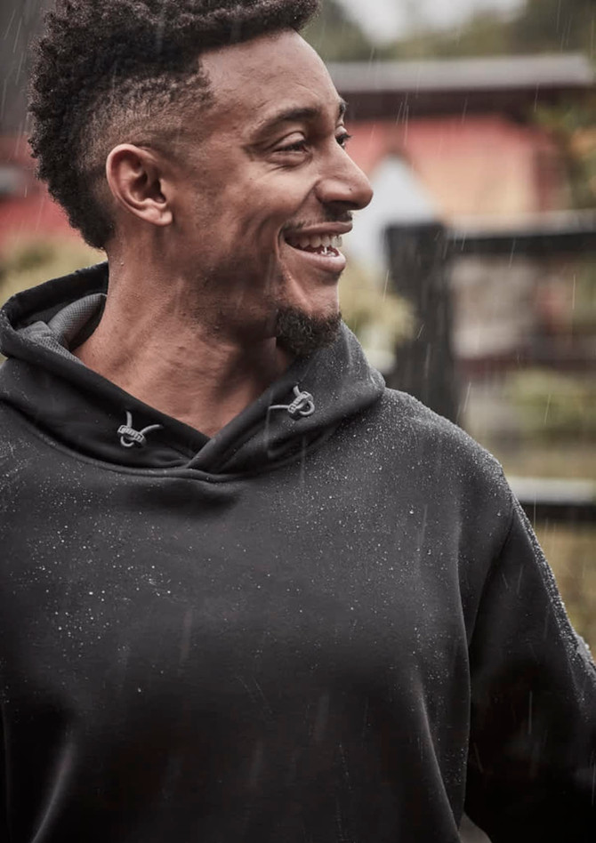A man wearing a black unisex water-resistant hoodie, smiling as rain falls around him. The hoodie features a logo.