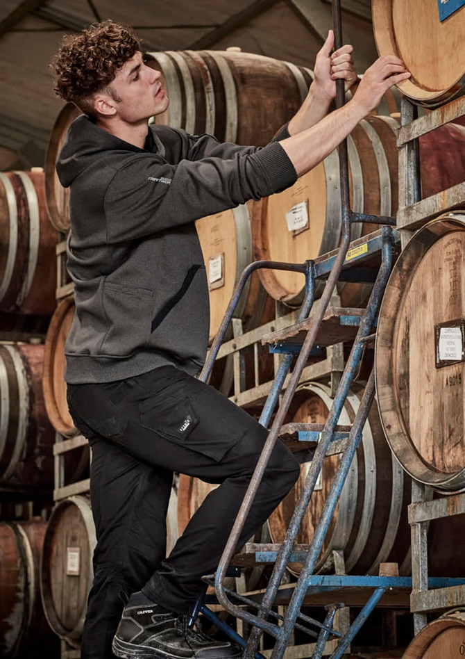 A person in a dark grey unisex water-resistant hoodie climbing a ladder among wine barrels. The hoodie has a logo.
