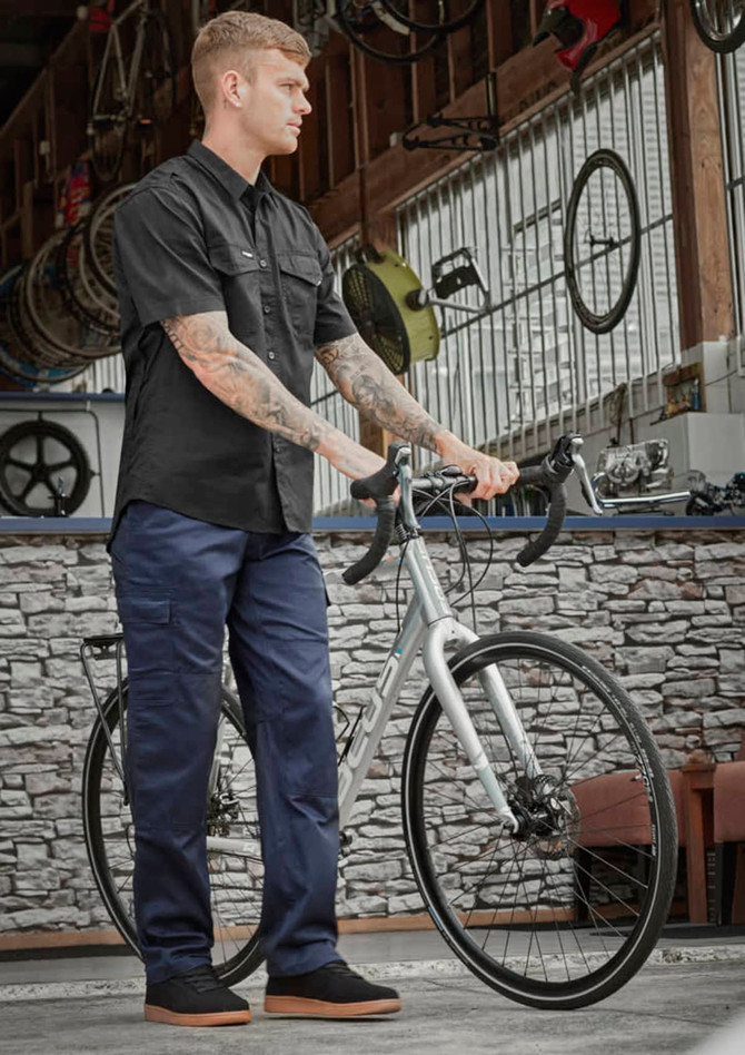 A person wearing navy midweight drill cargo pants and a black shirt stands beside a bicycle in a workshop setting.