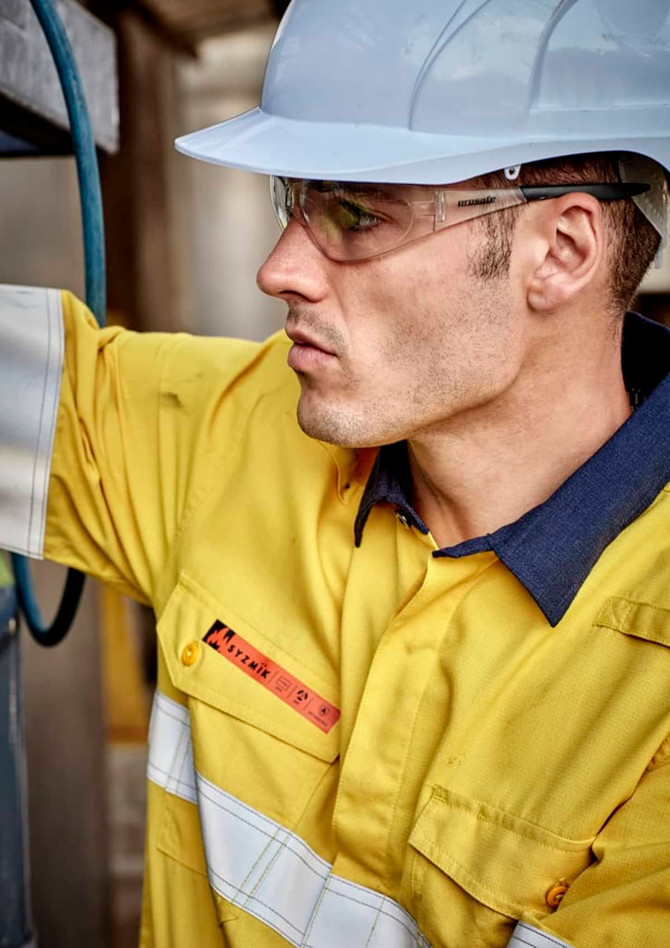 A man wearing a yellow work shirt with white reflective strips and a blue collar, accompanied by safety glasses and a hard hat.