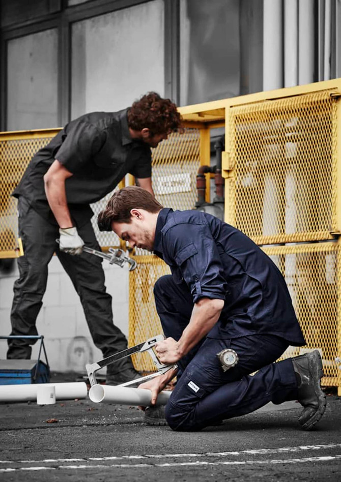 Two men in dark work shirts and pants engage in plumbing tasks outdoors near a yellow fence.
