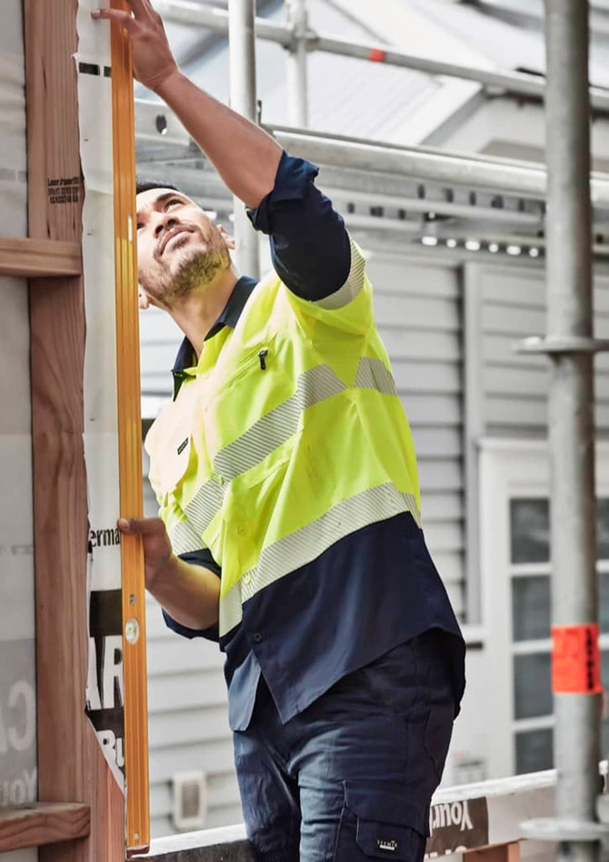A man wearing a hi-vis yellow and navy long sleeve work shirt examines a construction site using a level tool.