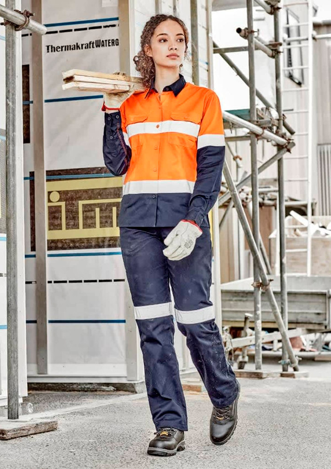 A woman in an orange and navy work shirt and matching pants stands on a construction site, holding wooden planks.