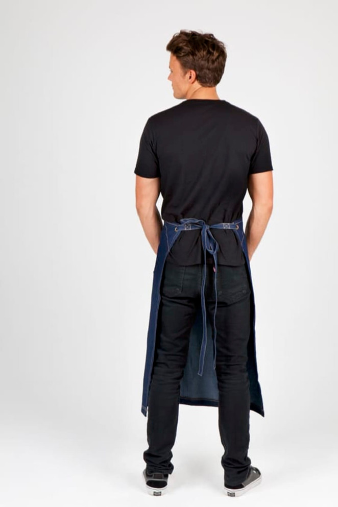A man wearing a long denim apron in navy blue, viewed from the back, paired with a black t-shirt and jeans.