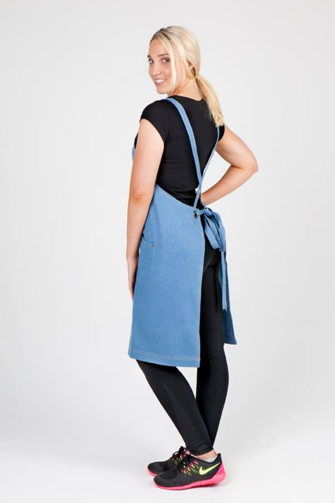 A blue full body cotton/denim apron with straps, featuring a model standing sideways in casual attire.