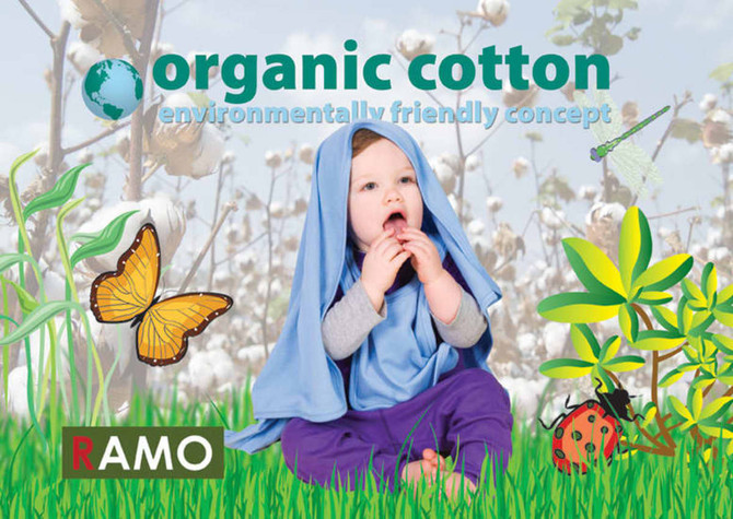 A baby in a blue blanket made of organic cotton, sitting on a green field with butterflies and plants in the background.