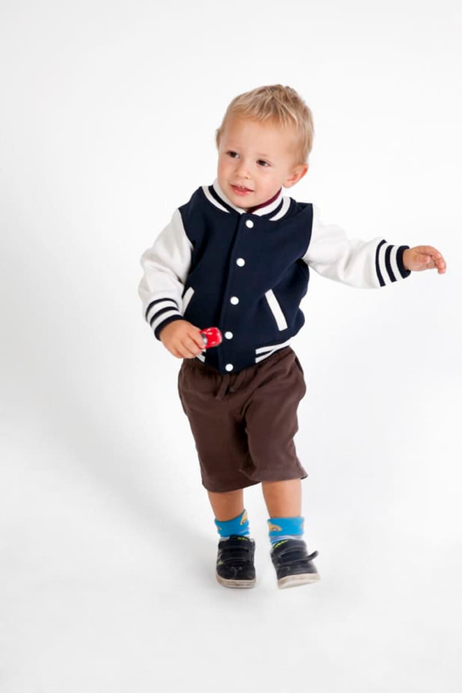 A child wearing a navy and white varsity jacket, brown shorts, and socks with blue accents, holding a small red object.