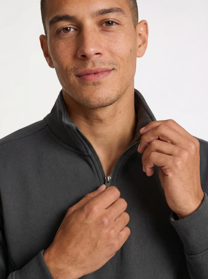 A man in a dark grey half-zip fleece jumper, slightly pulling at the collar. The jumper has a logo.