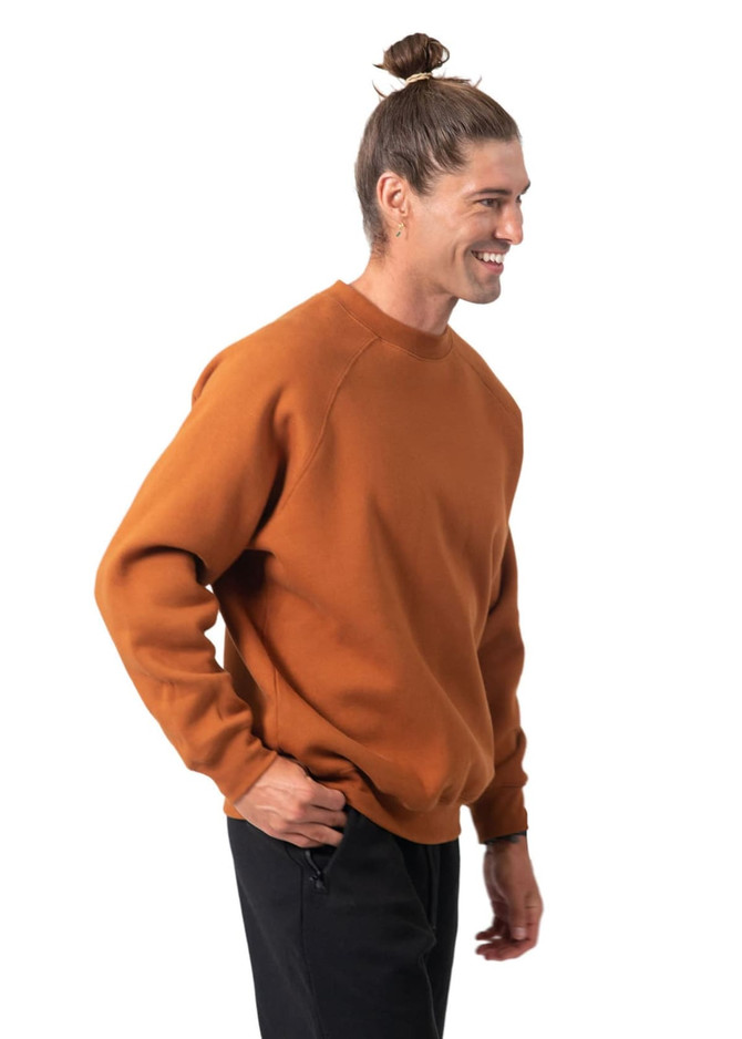 A man in a brown sweatshirt stands smiling, showcasing a casual style. The sweatshirt has a logo on it.
