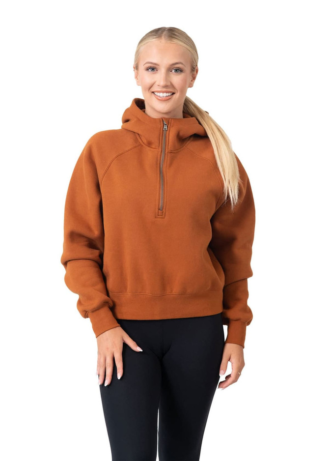 A woman wearing an orange half-zip jumper with a hood, paired with black leggings. The jumper has a logo.
