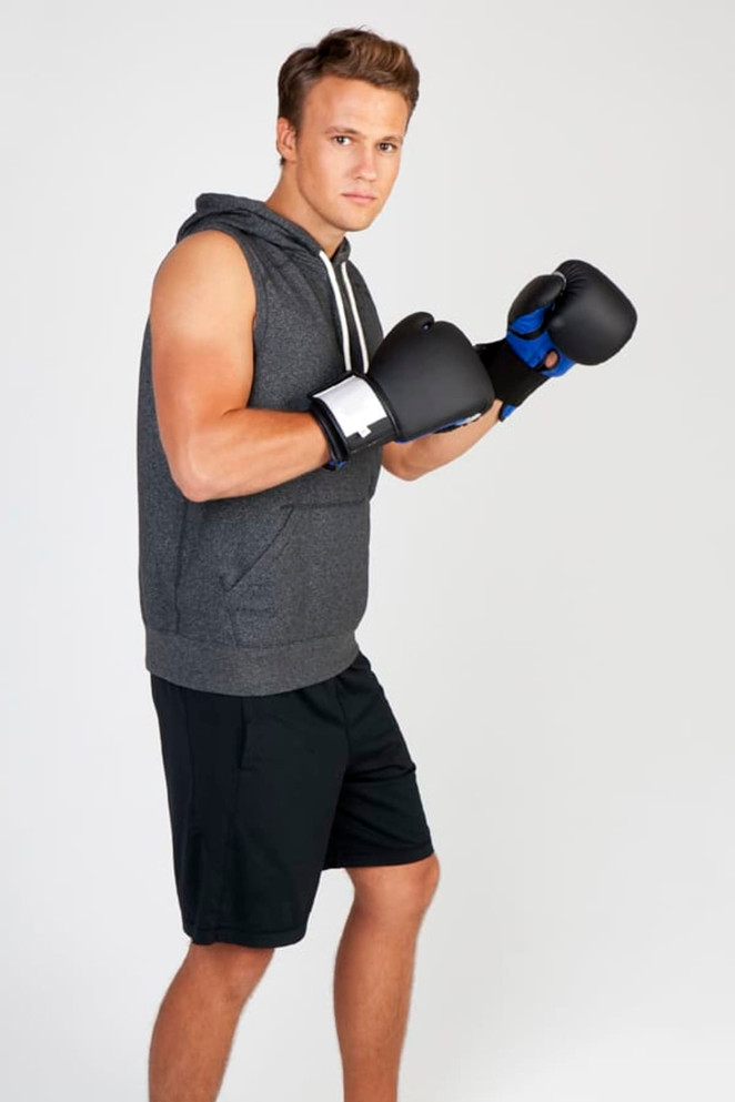 A man in a grey sleeveless hoodie and black shorts is posing with boxing gloves. The hoodie has a logo.