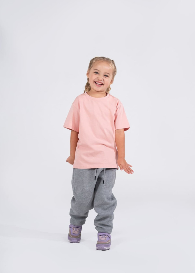 A child wearing a pink T-shirt and grey sweatpants, smiling in a simple, bright setting. The shirt features a logo.