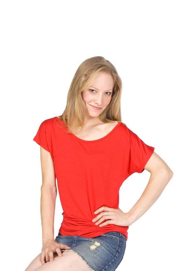 A woman sits on a stool, wearing a red bat wing T-shirt and denim shorts, paired with black high heels.