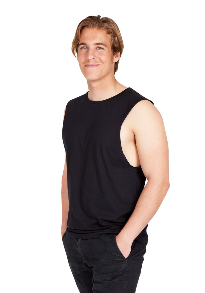 A man wearing a black combed cotton tank top, standing with hands in pockets and smiling. The tank has no sleeves and a relaxed fit.