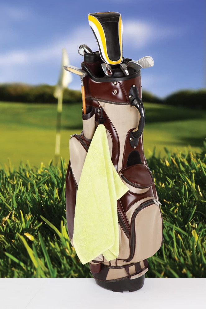 A bamboo golf towel in bright yellow hangs from a golf bag, surrounded by green grass and a blurred background of a golf course.