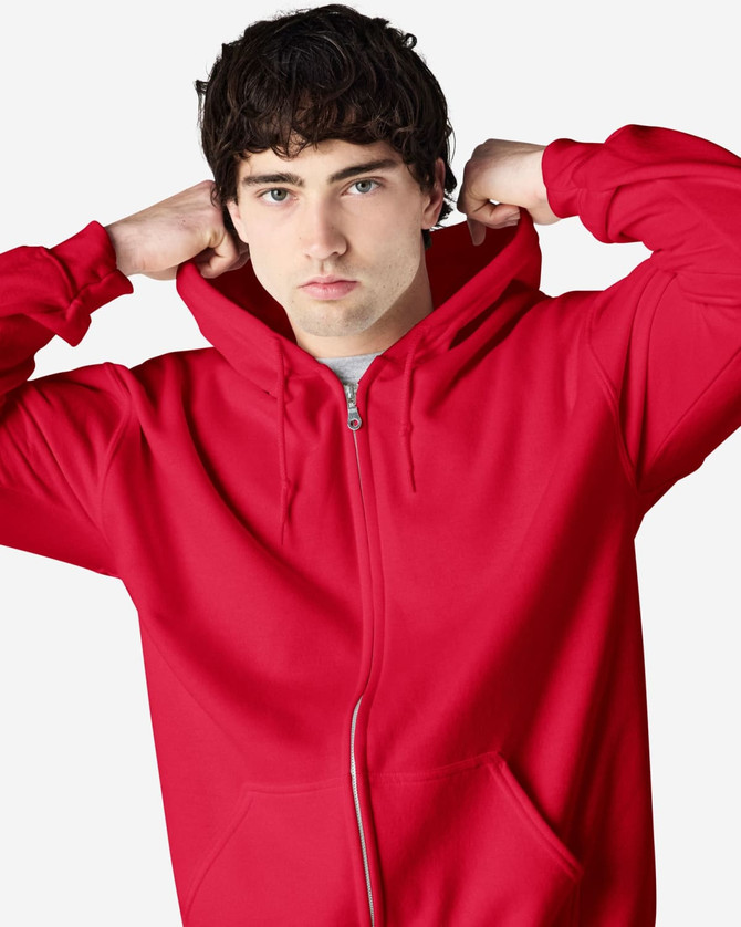 A red Gildan Heavy Blend adult full zip hooded sweatshirt on a model adjusting the hood. The sweatshirt features a logo.