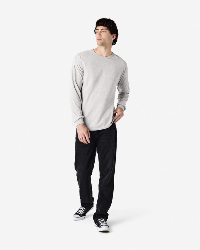 A long sleeve t-shirt in grey worn by a model, paired with black pants and sneakers, featuring a logo.
