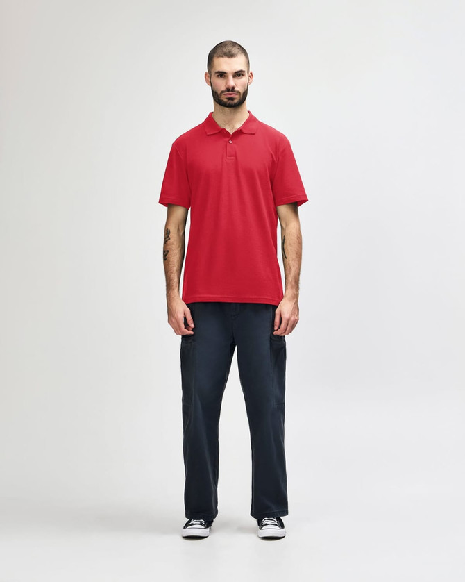 A red Gildan Softstyle adult double pique polo shirt displayed on a model wearing black jeans and sneakers. The shirt has a logo.