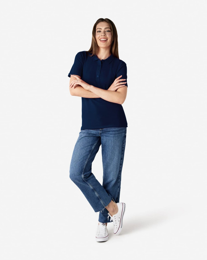 A woman stands smiling in a navy blue Gildan Softstyle ladies' polo shirt and denim jeans, wearing white sneakers.
