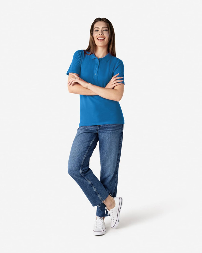 A woman poses in a bright blue polo shirt and jeans, standing confidently with arms crossed. The polo features a logo.