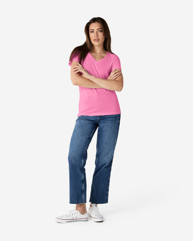 A woman stands wearing a pink Gildan Softstyle ladies' V-neck T-shirt and blue jeans, with white sneakers.