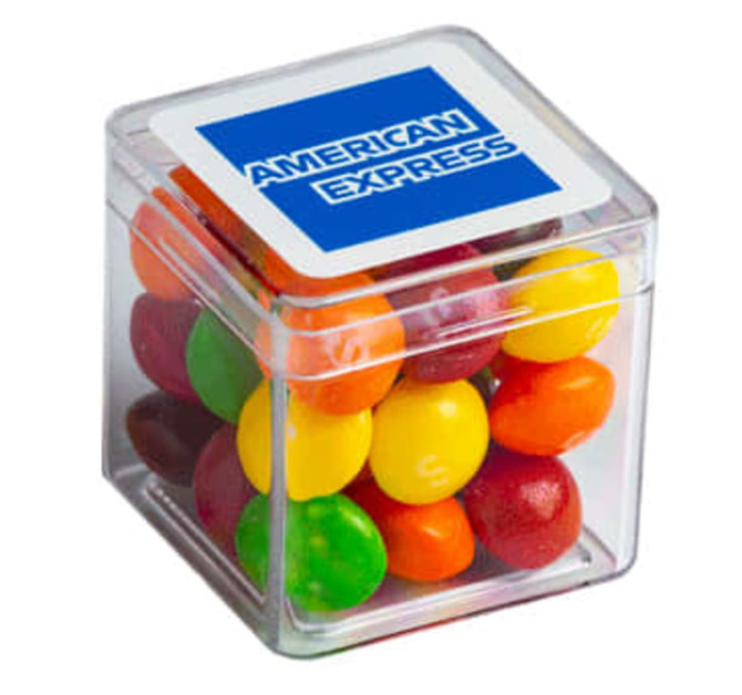 A transparent cube containing colourful Skittles in various shades, with a branding logo on the lid.