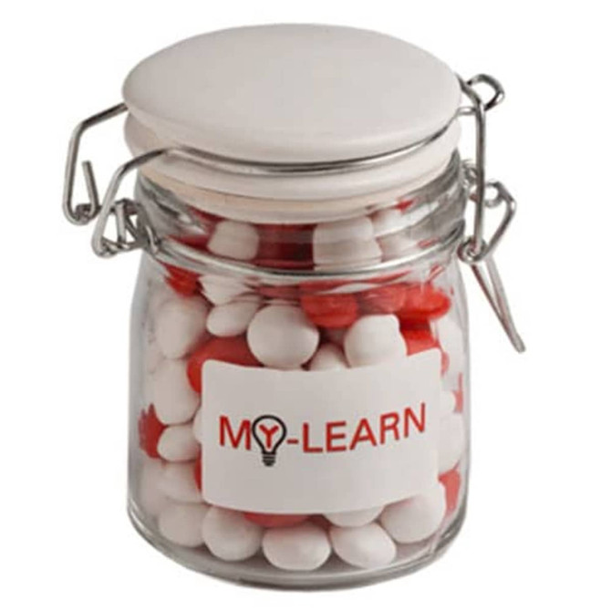 A glass clip lock jar with a white lid, filled with red and white chewy fruit sweets. The jar features a logo.