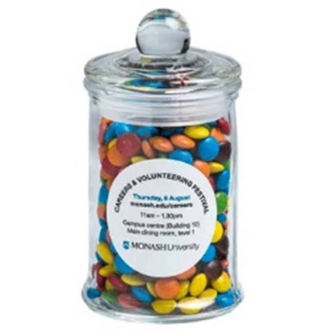 A small glass apothecary jar filled with colourful chocolate beans, featuring a round lid and a logo.