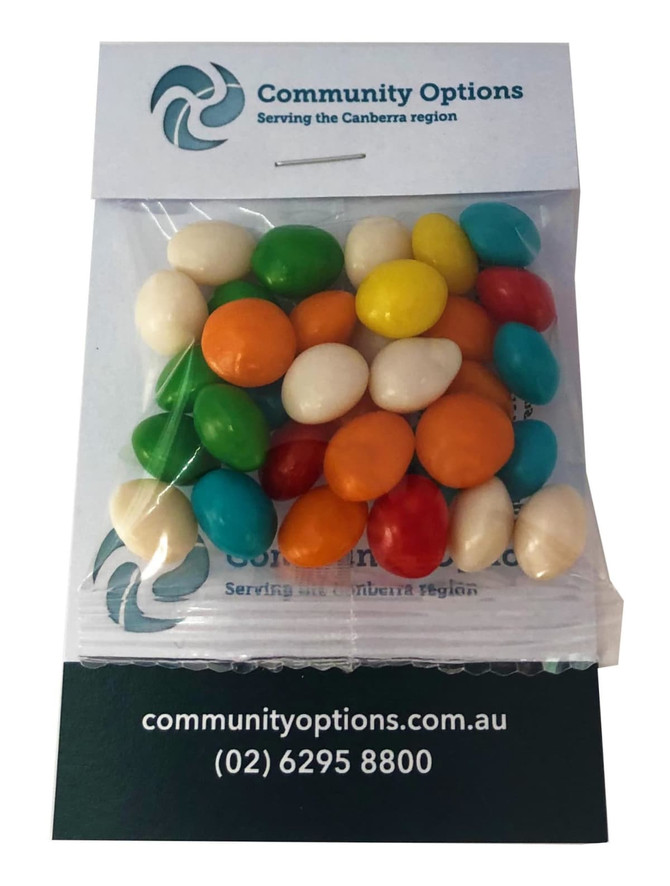 A clear bag filled with colourful chewy fruit candies, including green, orange, yellow, blue, and white pieces, placed on a card with a logo.