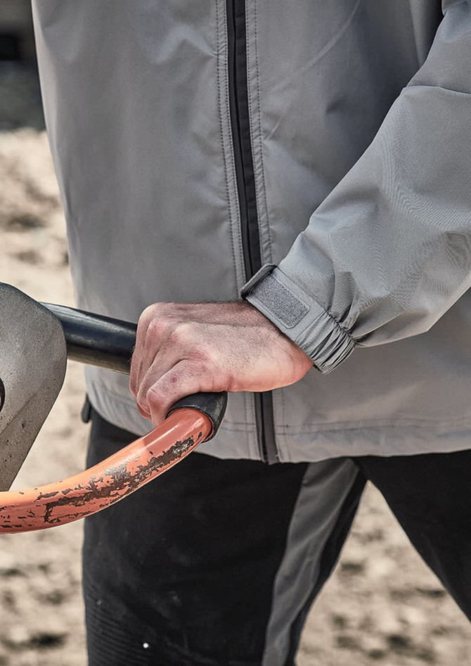 A close-up of a grey unisex packable rain jacket, focusing on the cuff and hand gripping a tool handle.