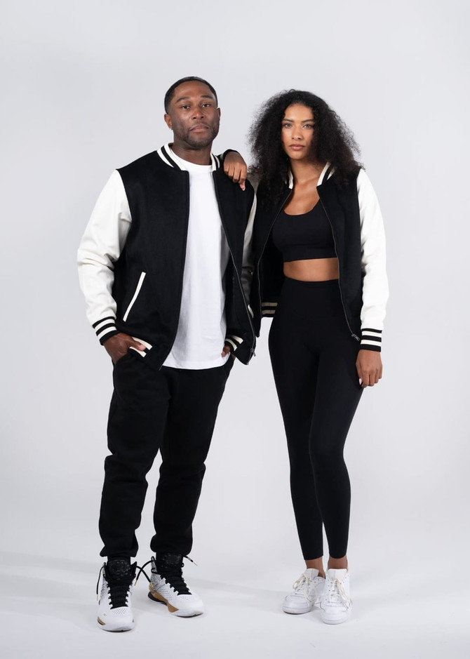 A black and cream varsity jacket featuring a logo, displayed on a man and a woman in casual attire.