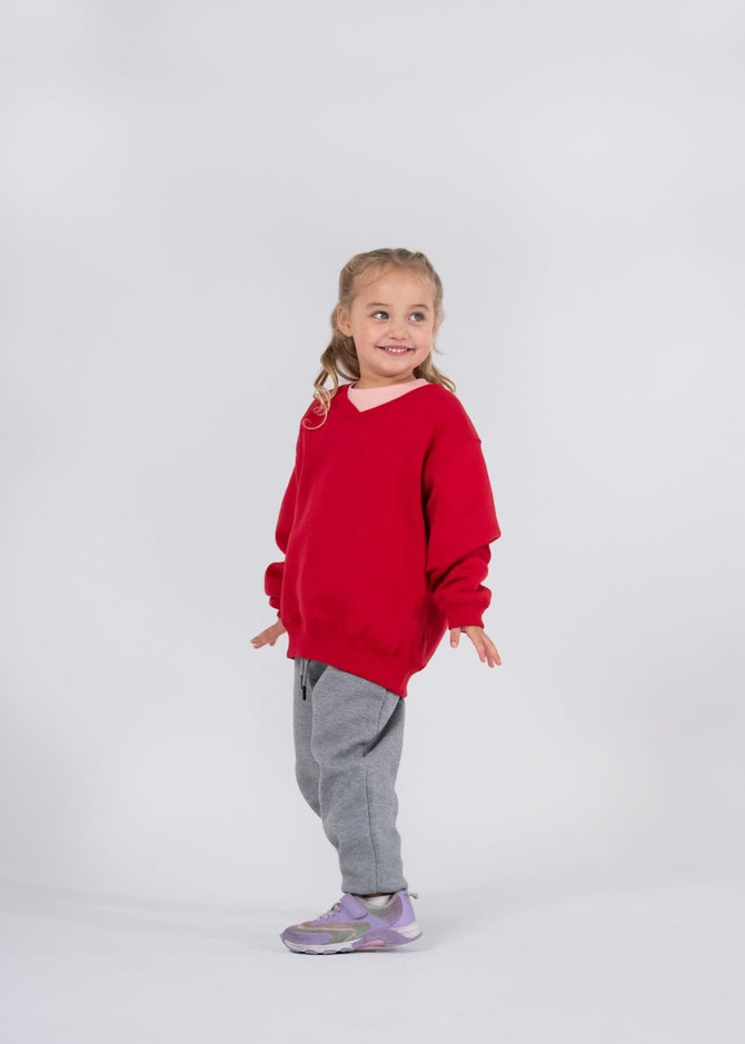 A cute child in a red fleece jumper and grey pants, smiling and standing in a playful pose.